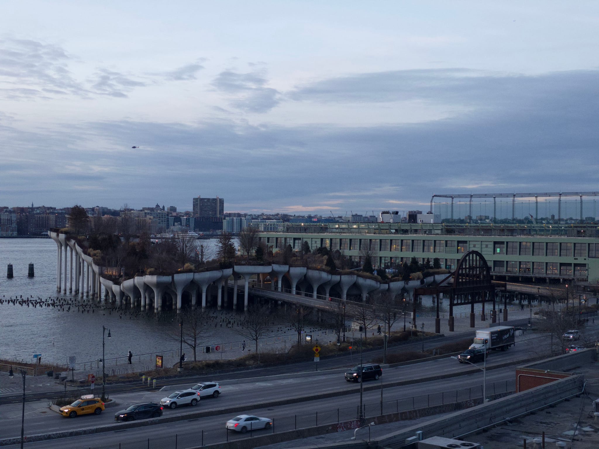 A waterfront park with a unique pier structure extends into the water, surrounded by a cityscape under a cloudy sky. A road with vehicles runs alongside the park
