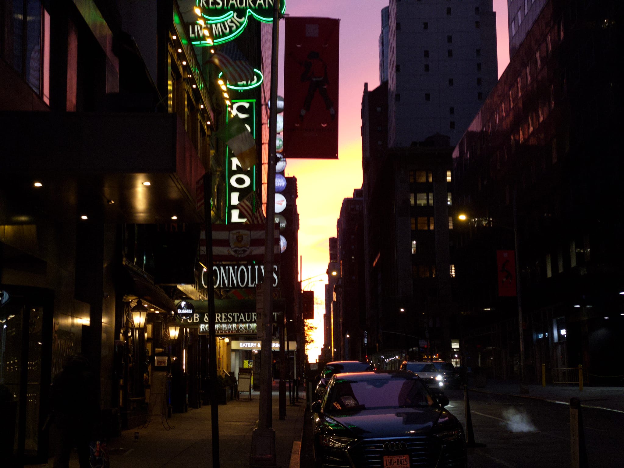 A city street at dusk with neon signs, parked cars, and tall buildings silhouetted against a colorful sky