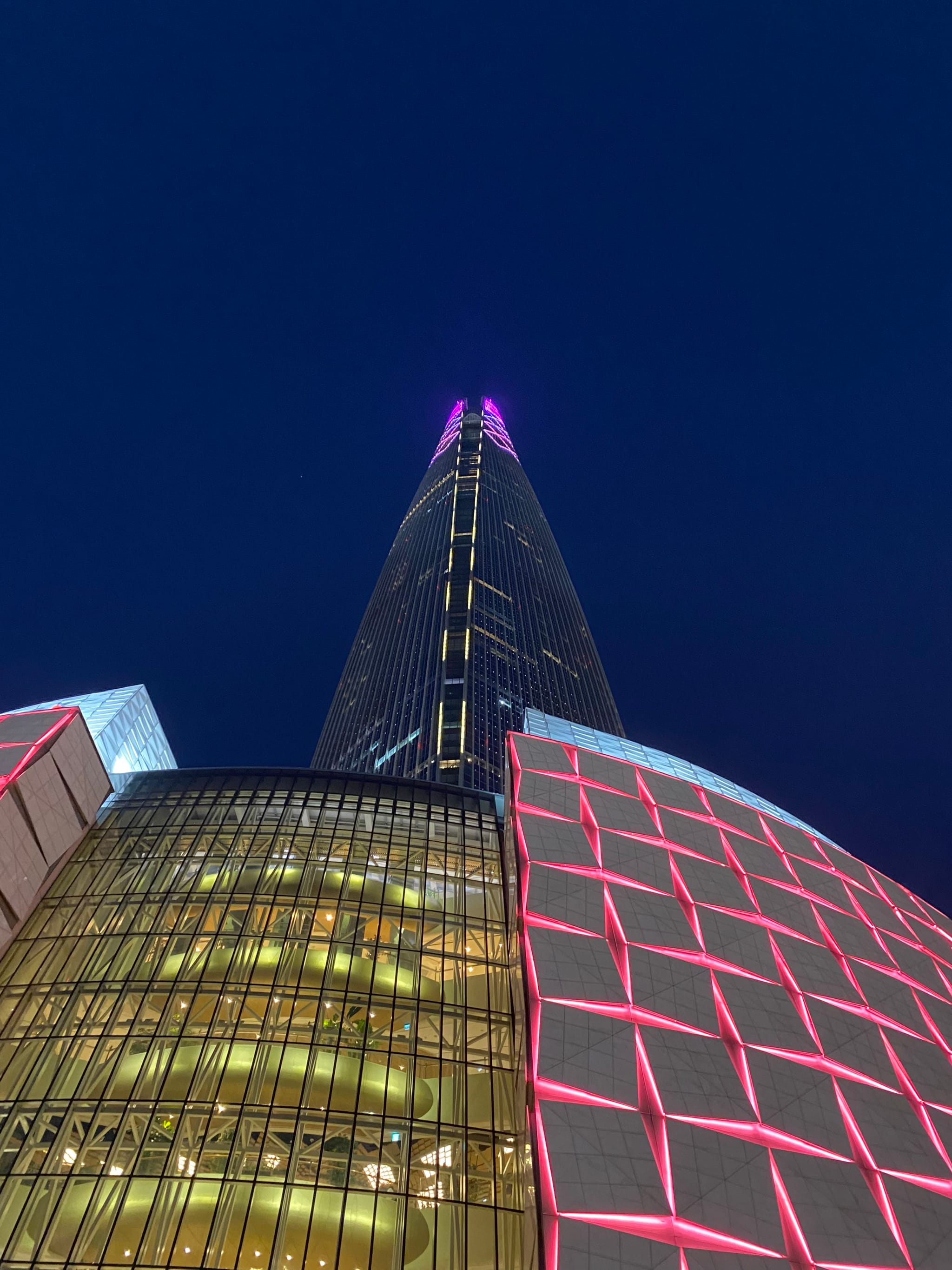 A tall, illuminated skyscraper with a pointed top against a dark blue sky, featuring a grid pattern with pink lighting on the lower sections