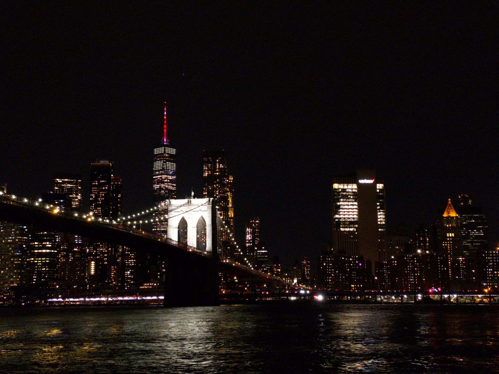 A nighttime cityscape featuring a brightly lit bridge and illuminated skyscrapers reflecting on the water