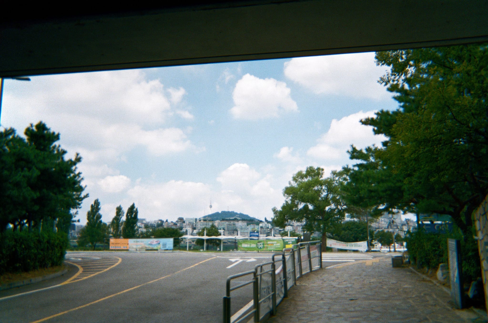 Paved walkway beside a road with railings and trees leading toward a distant bridge along a river under a partly cloudy sky