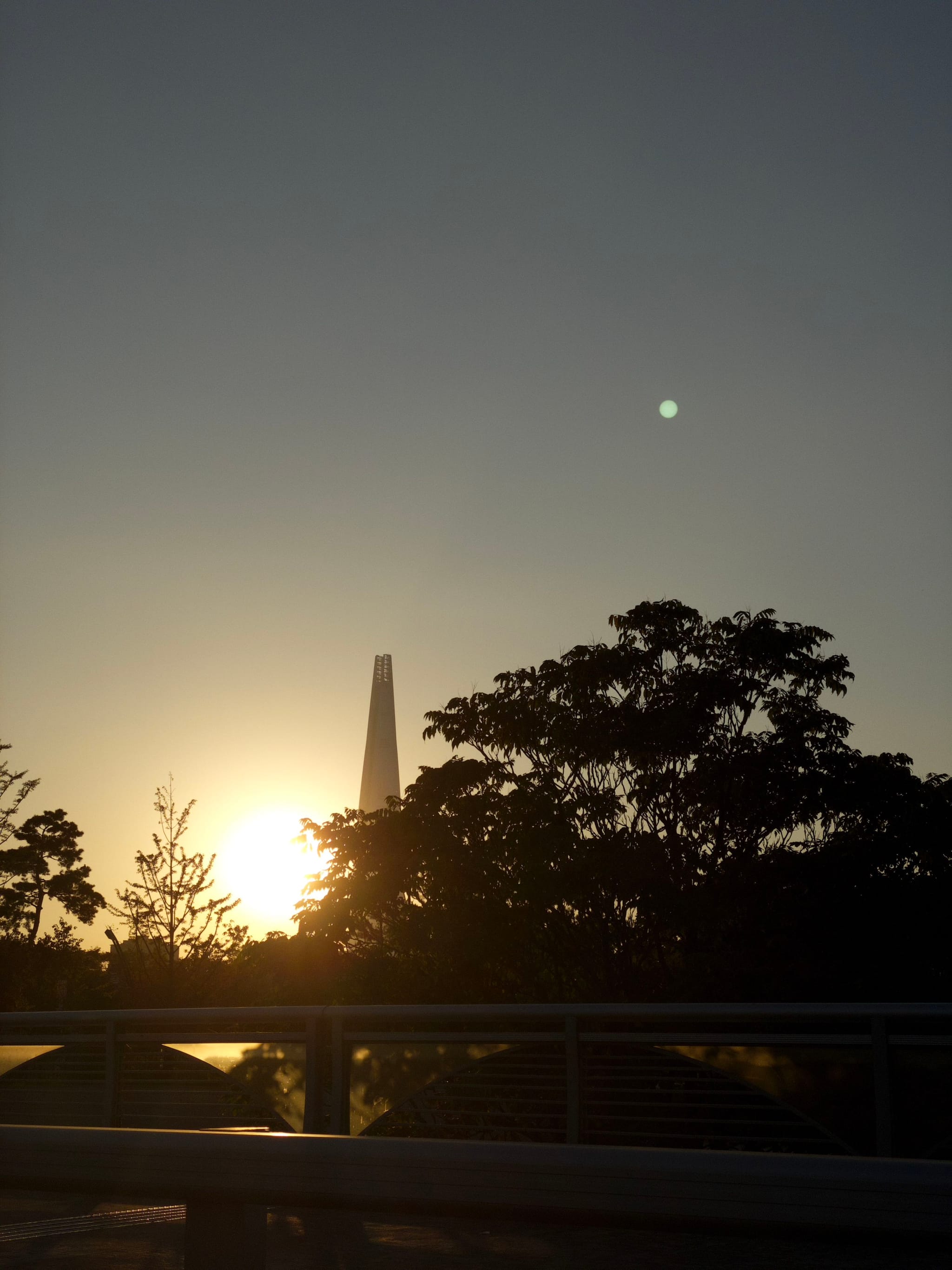 A sunset scene with the sun partially hidden behind trees, a tall structure in the background, and a railing in the foreground