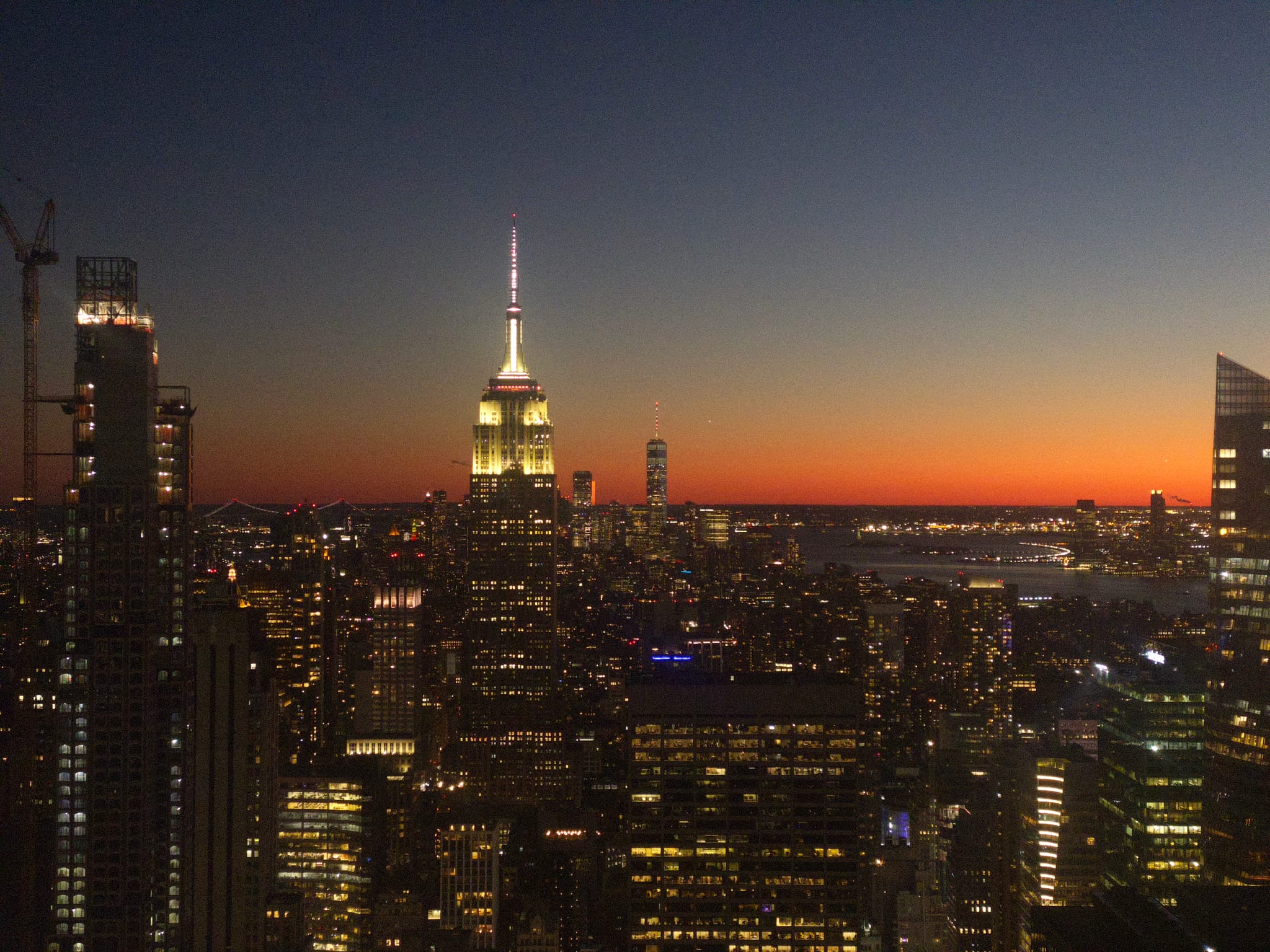 A city skyline at dusk with illuminated skyscrapers, including a prominent tower with a spire, set against a vibrant sunset