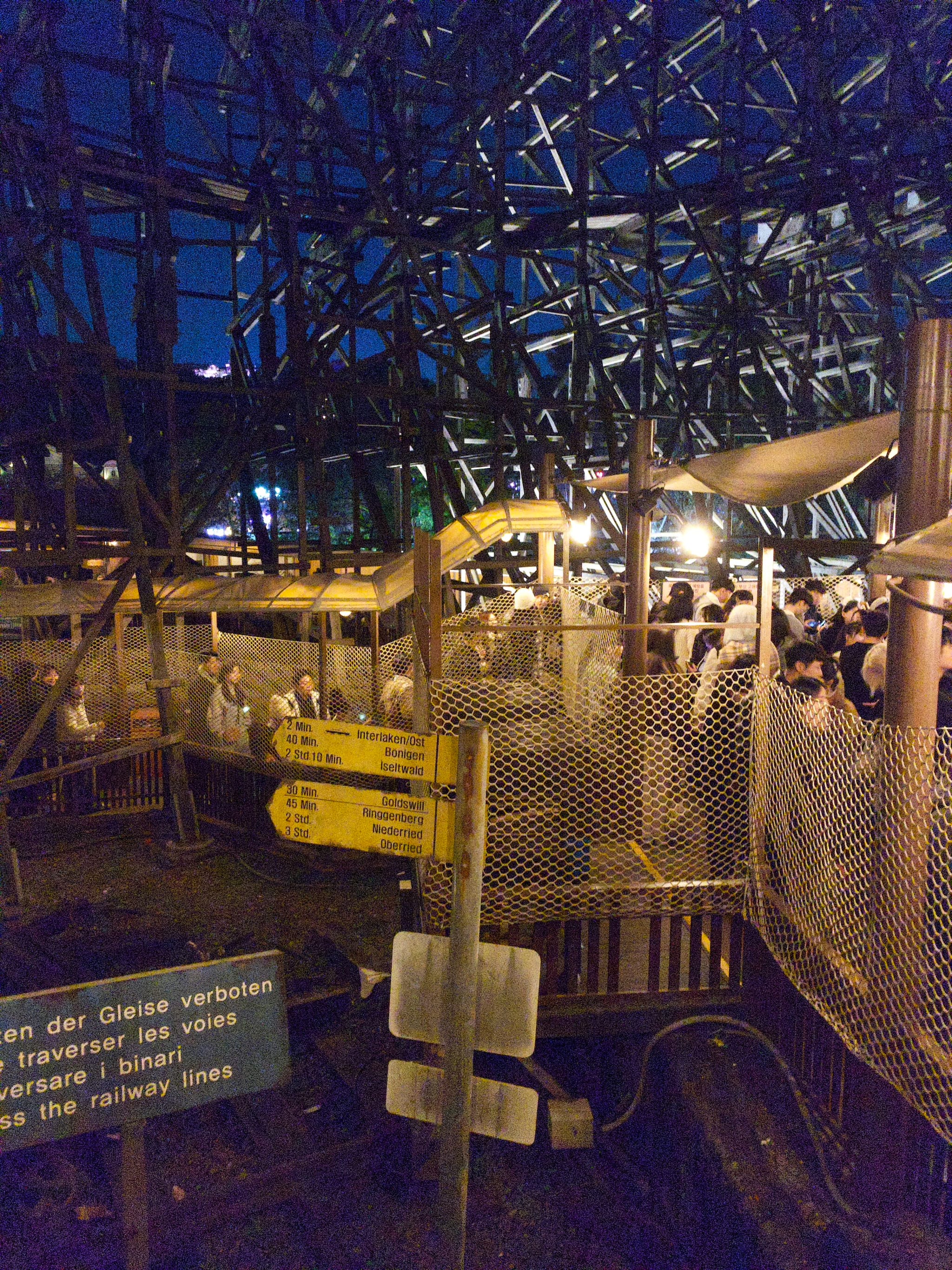 A nighttime amusement park scene with a wooden roller coaster structure, illuminated by lights. A crowd of people is visible in the queue area, surrounded by safety netting and directional signs