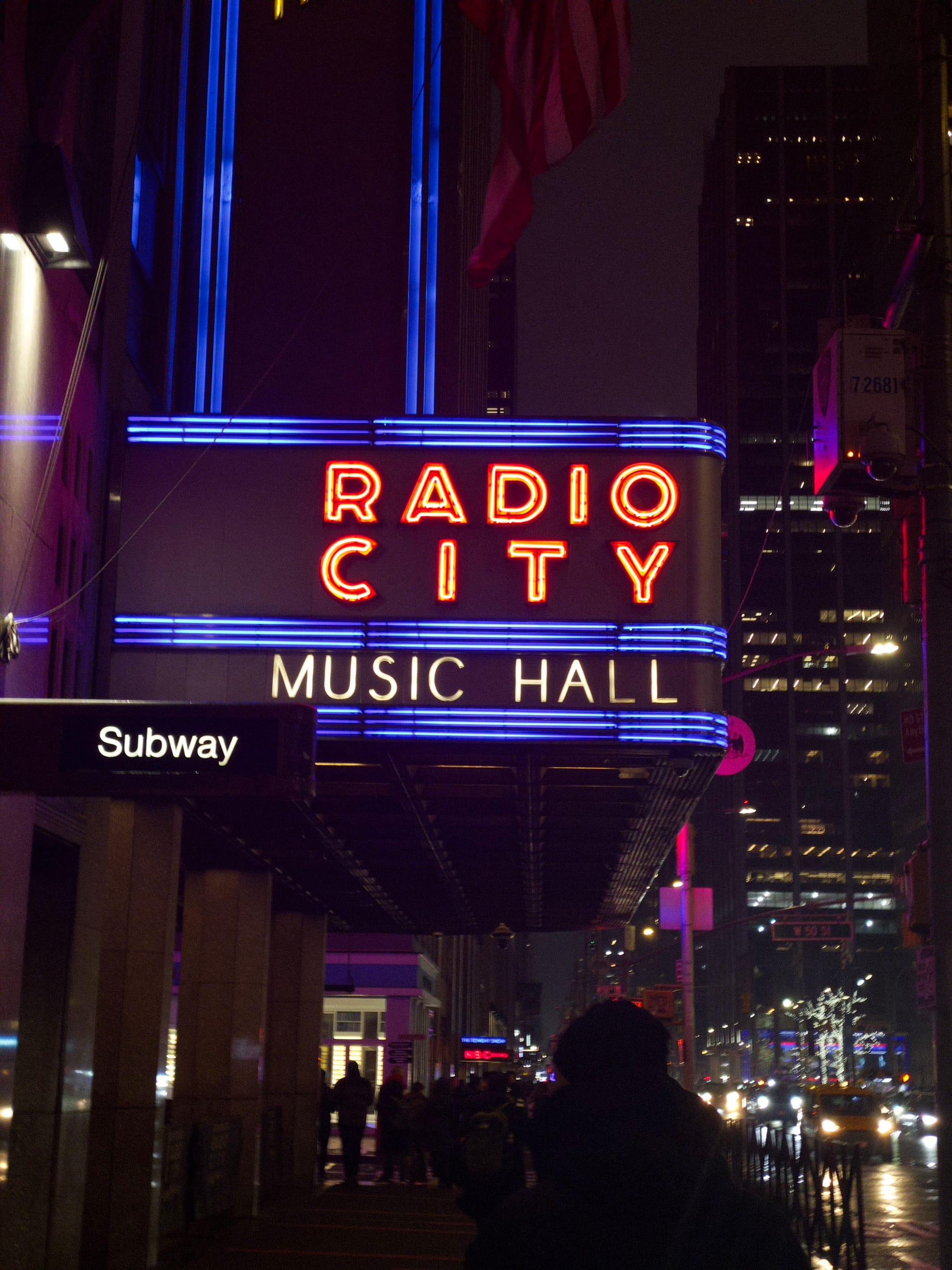 A neon sign for Radio City Music Hall at night, with surrounding city lights and a subway entrance visible