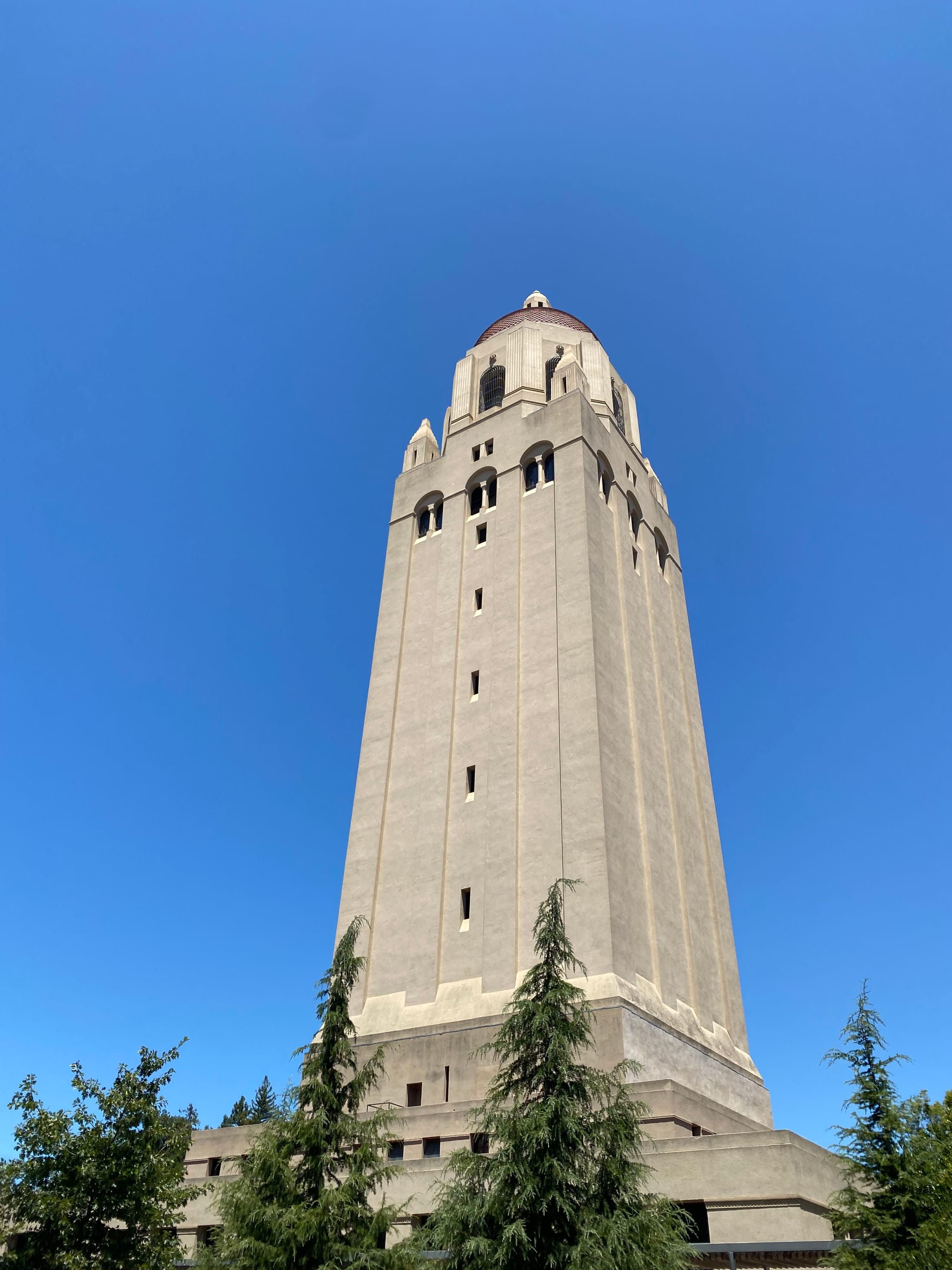 A tall, beige tower with a red-tiled roof set against a clear blue sky, surrounded by trees at the base