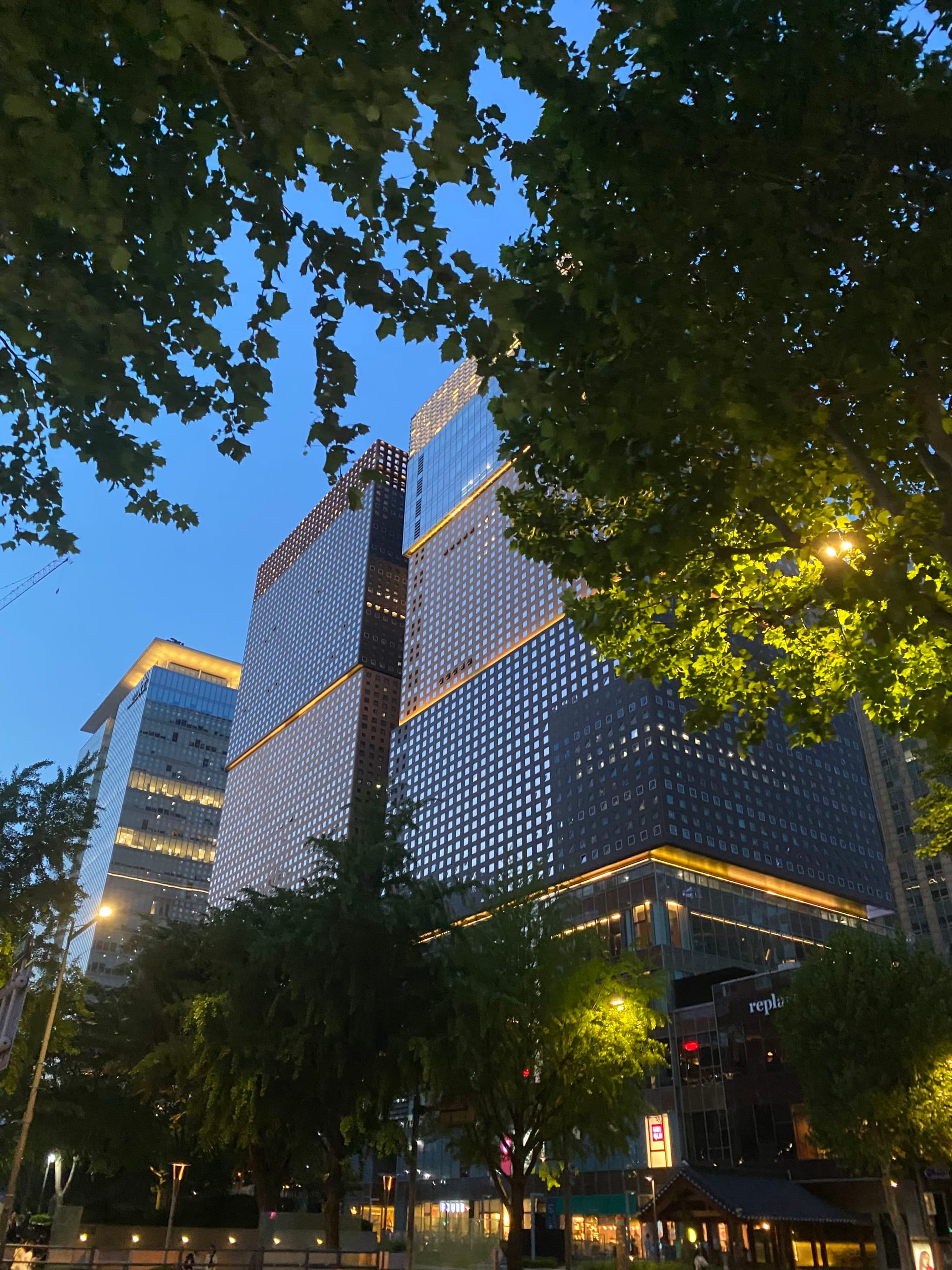 A modern cityscape with tall, illuminated buildings surrounded by trees, set against a twilight sky
