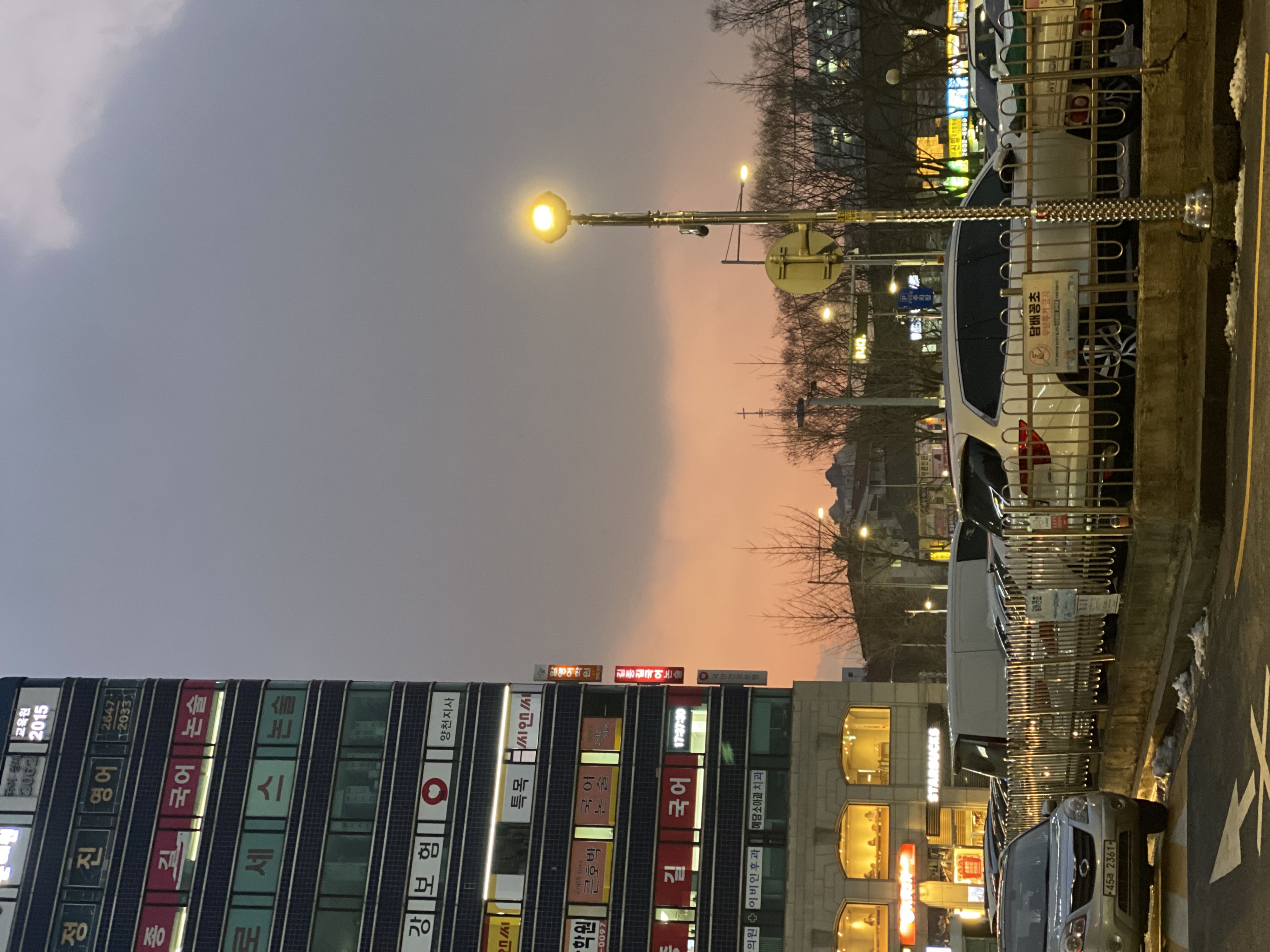 A cityscape at dusk with a tall building on the left, a parking lot with cars in the foreground, and a streetlamp illuminating the scene. The sky is cloudy with a hint of sunset colors near the horizon