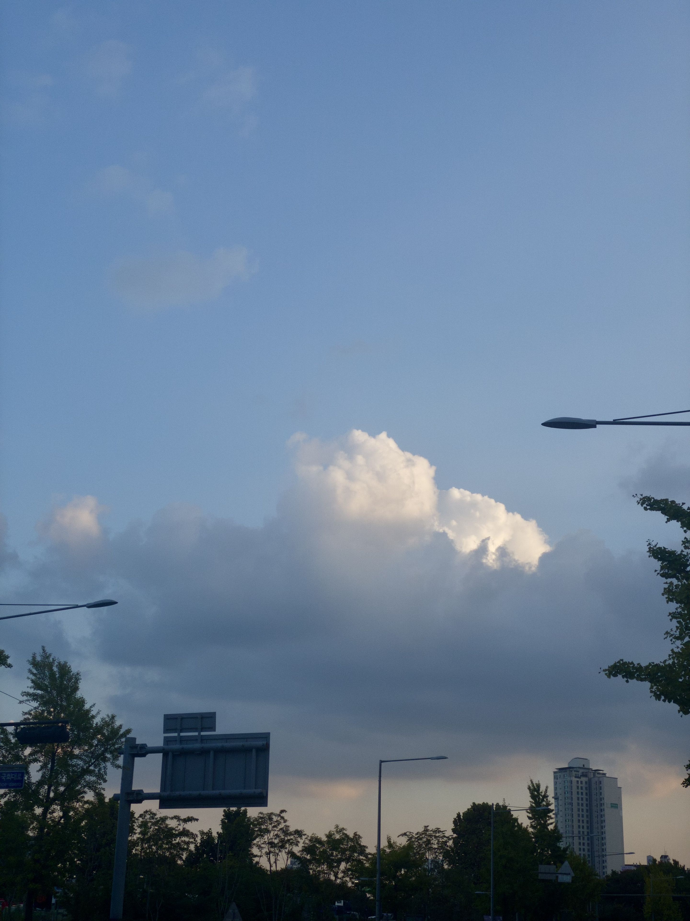A cityscape with a clear blue sky and fluffy clouds, featuring streetlights, trees, and a tall building in the background