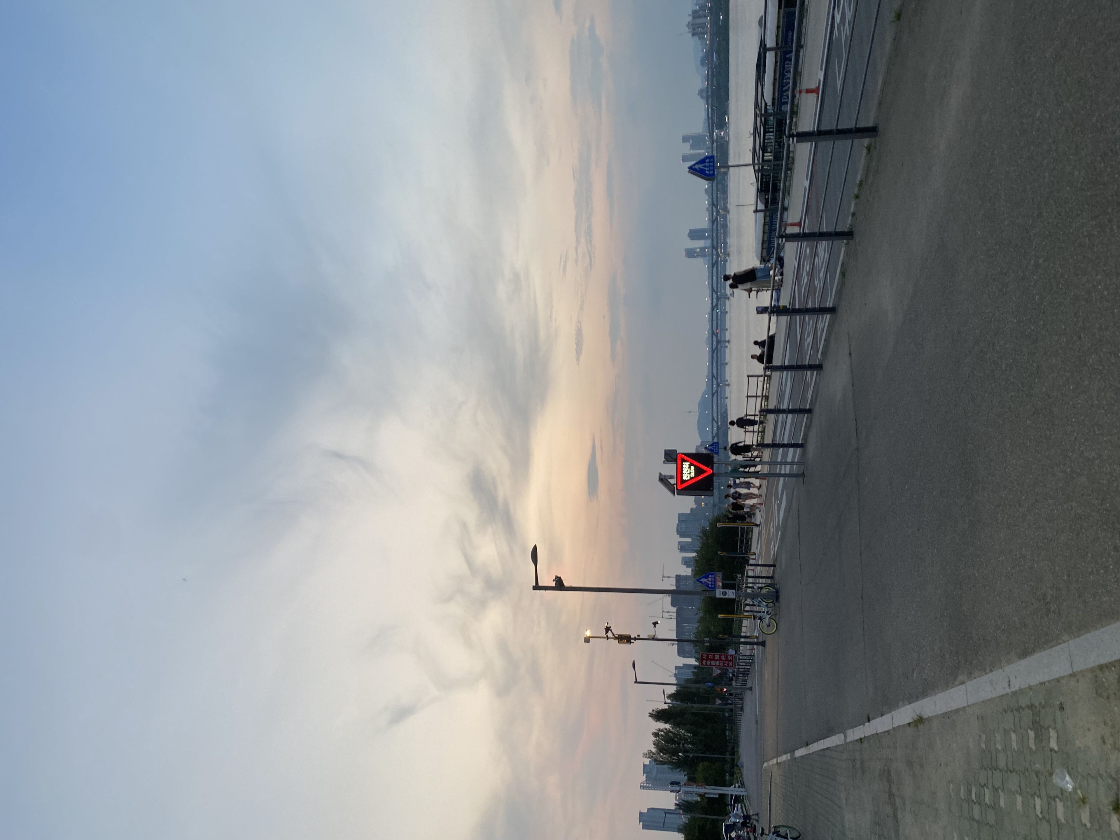 A riverside walkway at dusk with a few people strolling, streetlights, and a distant city skyline under a cloudy sky