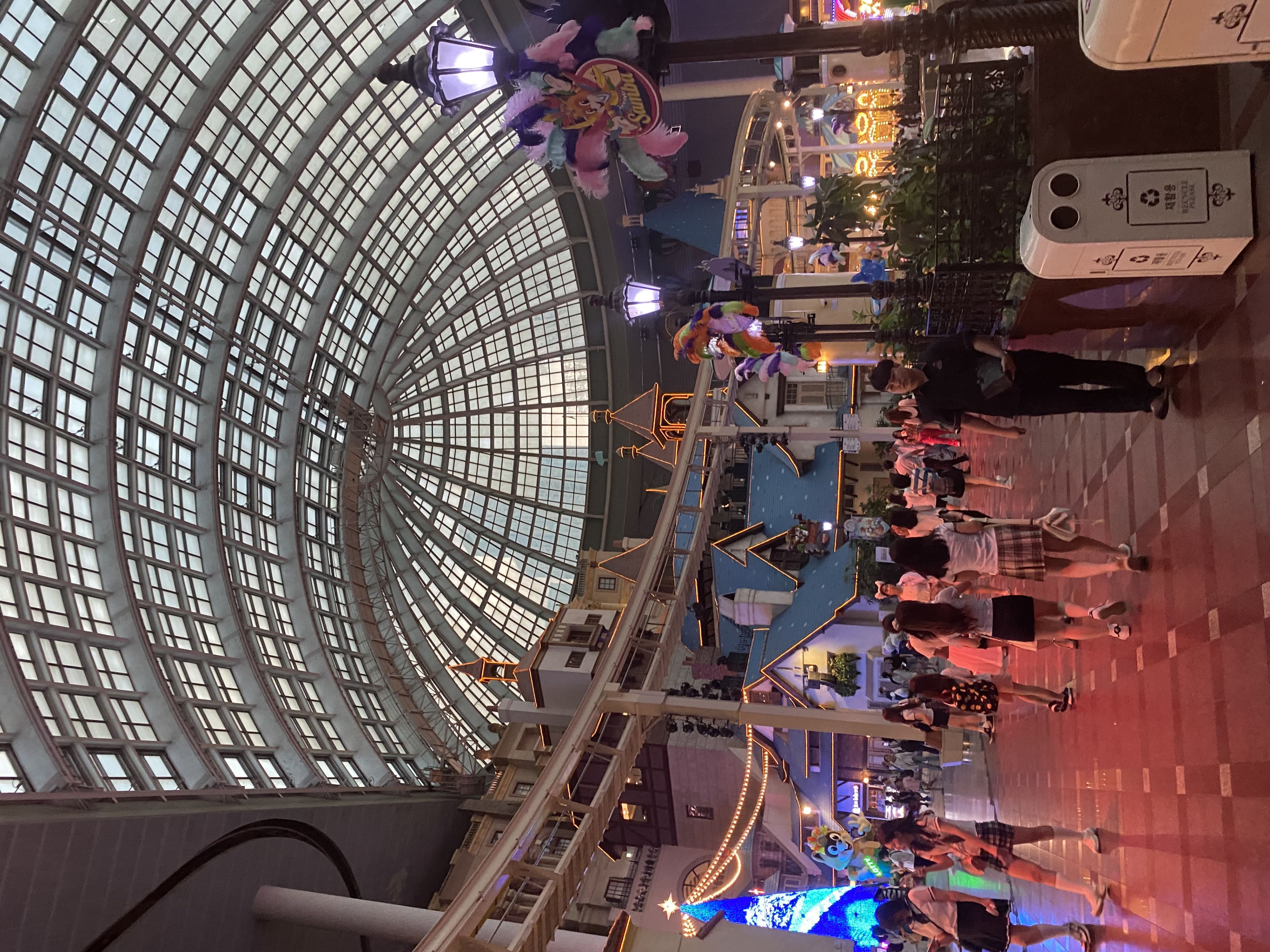 An indoor amusement park with a large glass dome ceiling, colorful decorations, and people walking around