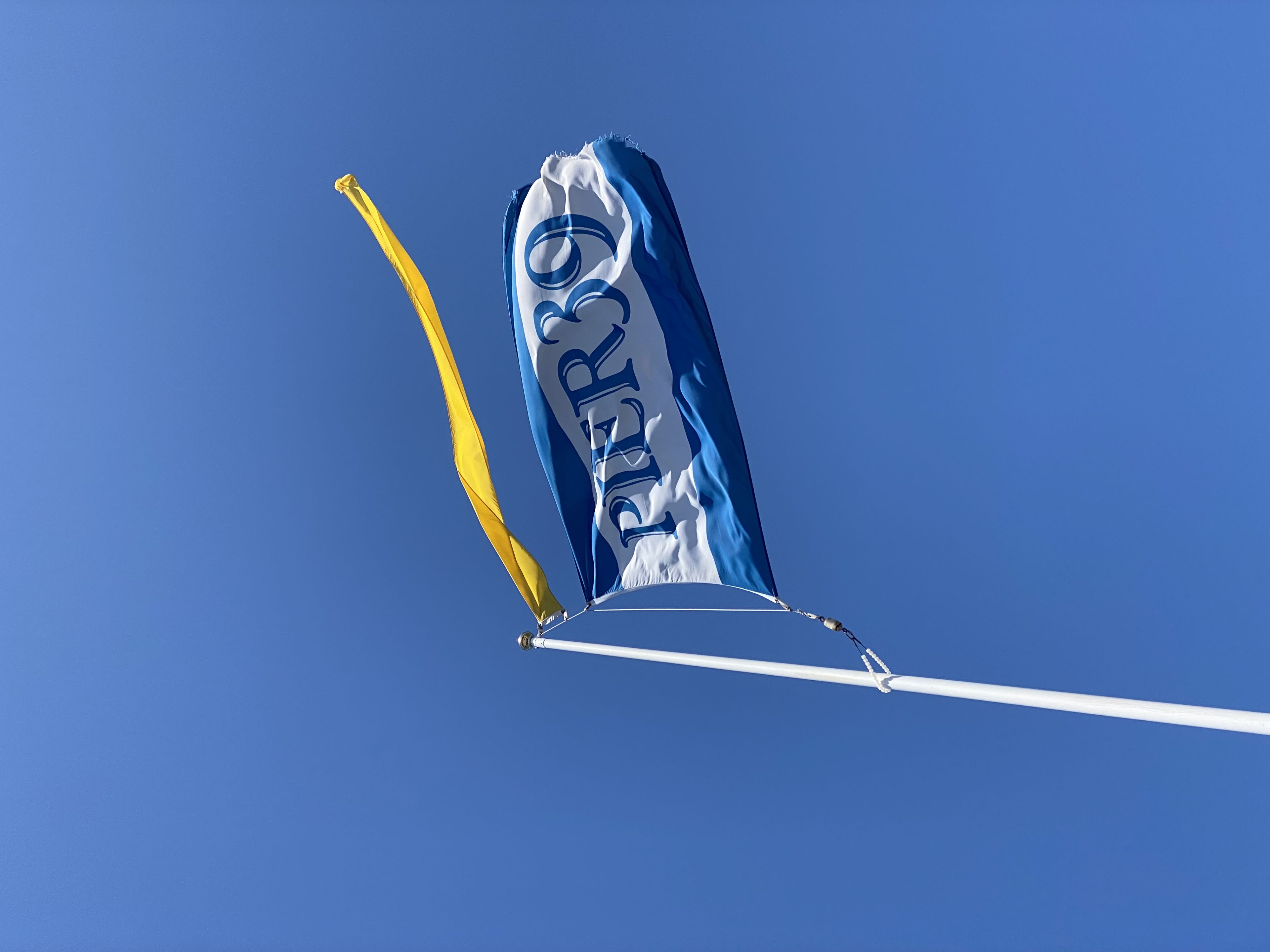 A blue flag with the word PIER 39 flutters on a pole against a clear blue sky, accompanied by a yellow pennant
