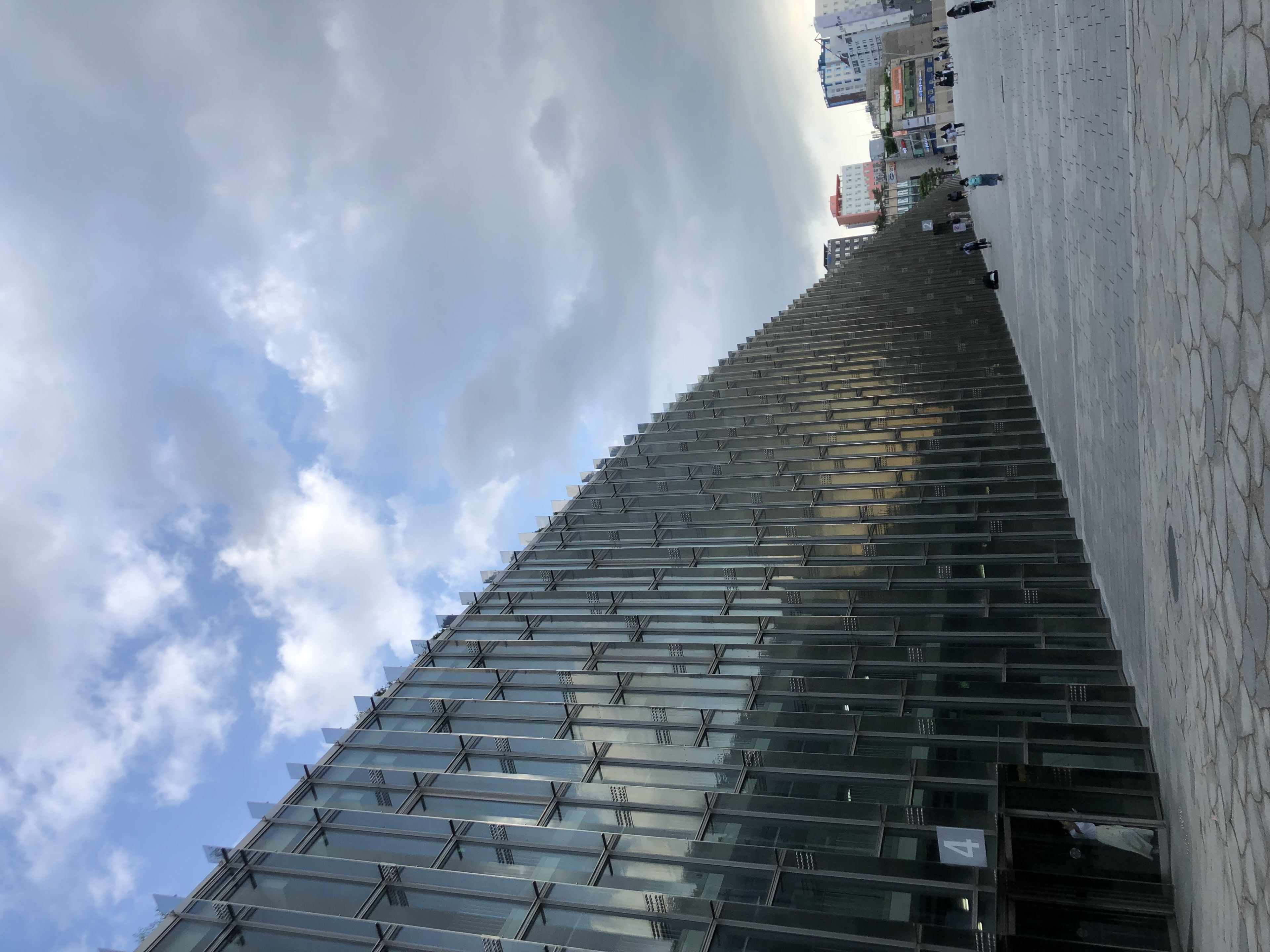 A modern glass building with a reflective facade, set against a partly cloudy sky, with a paved walkway and a few people in the distance