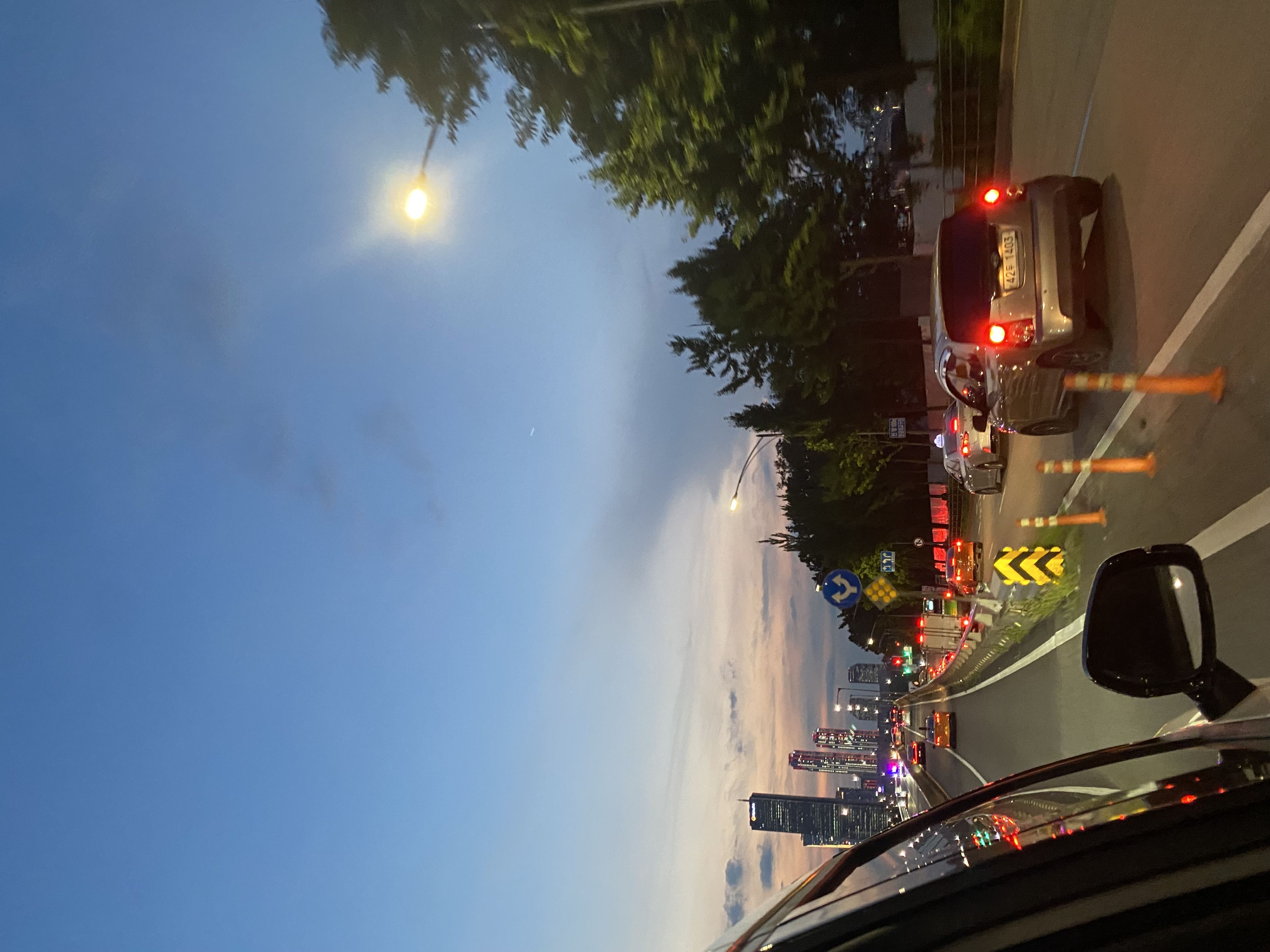 A city street at dusk with cars driving along a road lined with traffic cones. The sky is a gradient of blue and orange, and streetlights are illuminated. Trees and buildings are visible in the background
