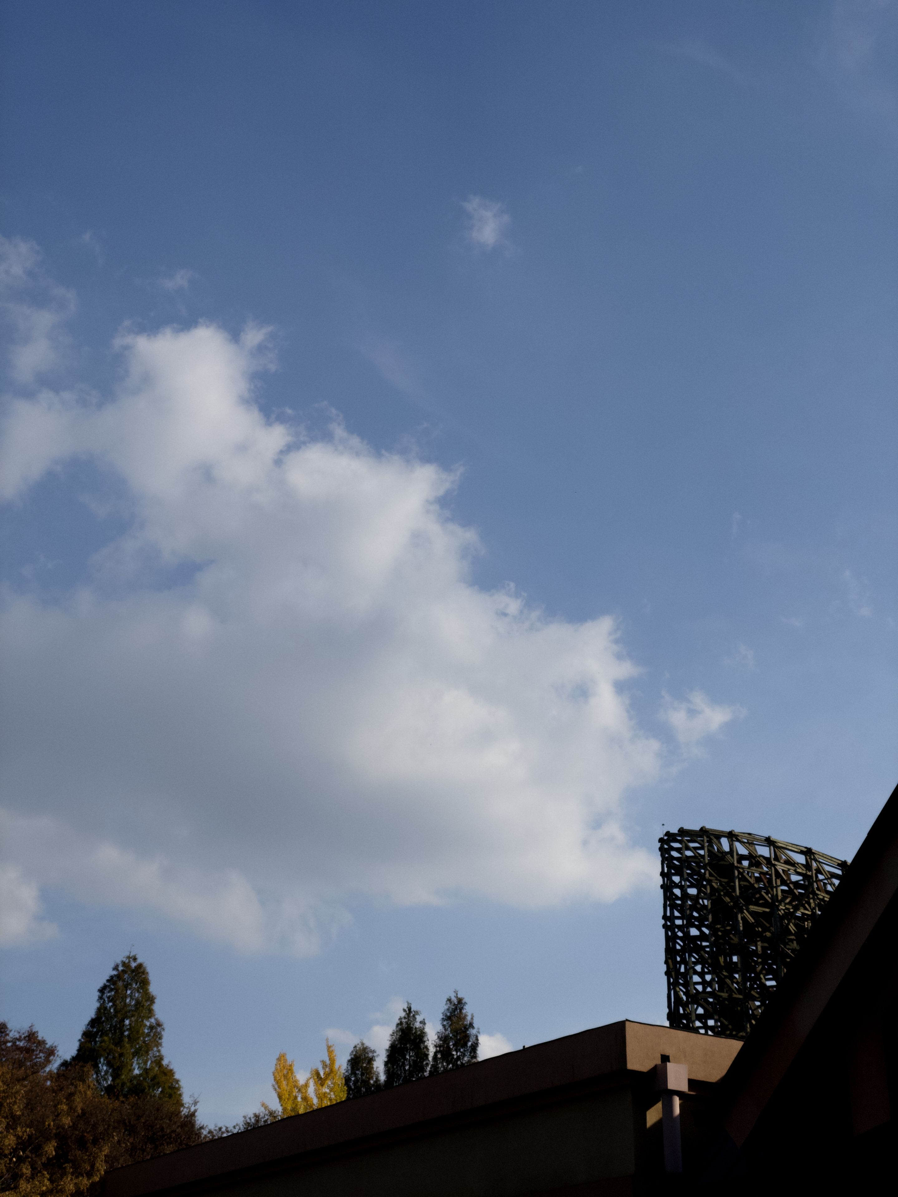 A clear blue sky with scattered clouds above a building, featuring a rooftop with a decorative metal structure and trees in the background
