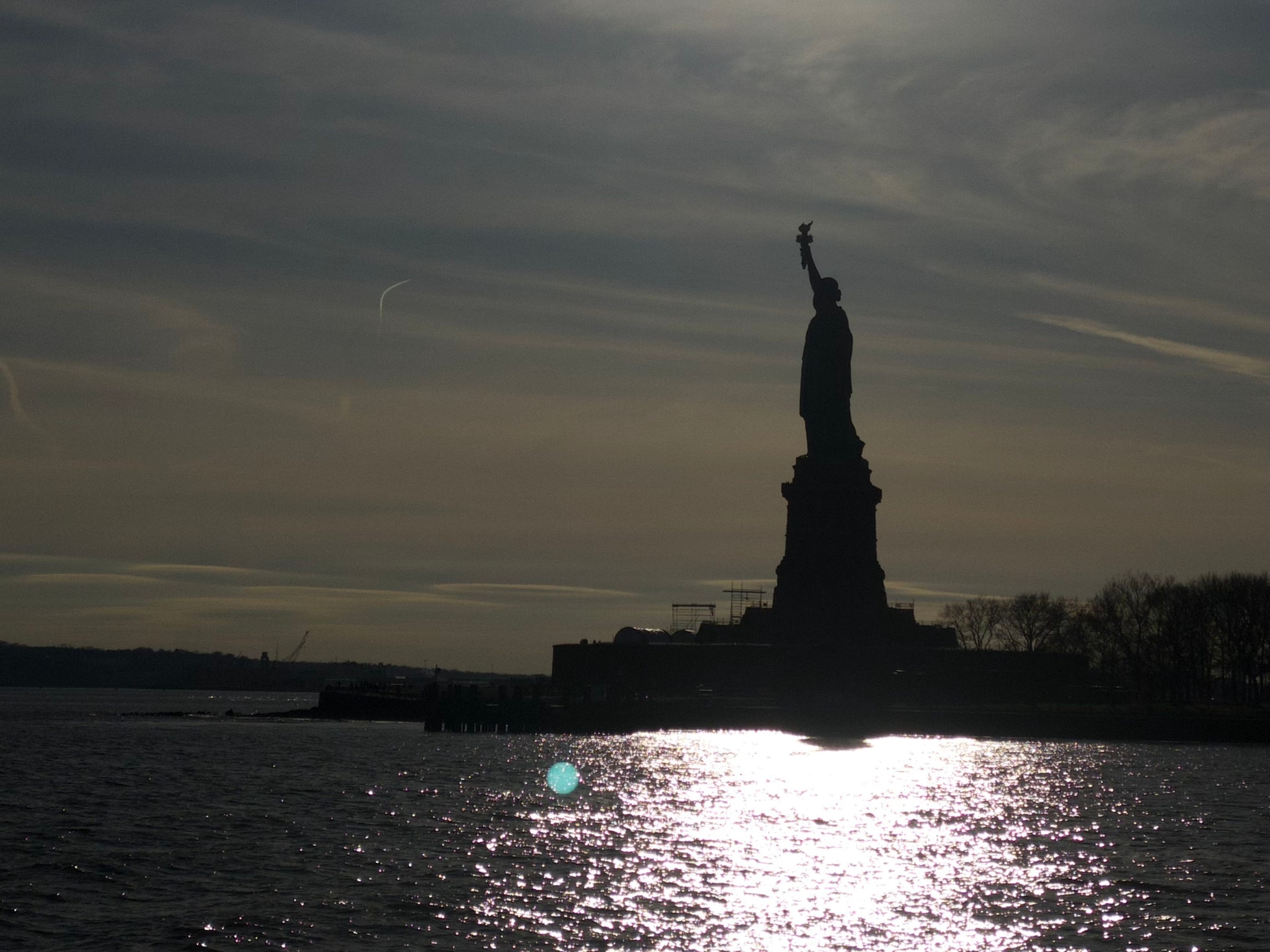 A silhouette of the Statue of Liberty against a darkening sky, with sunlight reflecting on the water