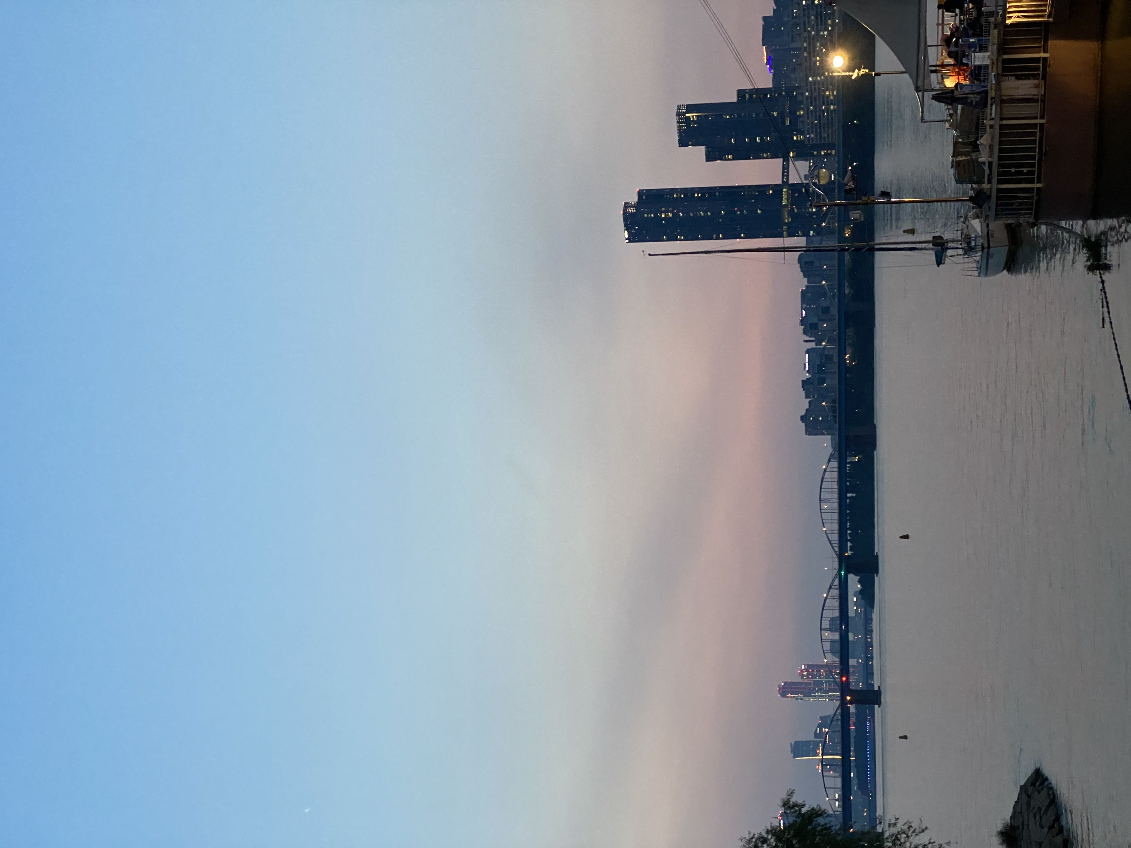 A waterfront cityscape at dusk with tall buildings, a bridge, and boats docked along the shore under a clear sky transitioning from blue to pink