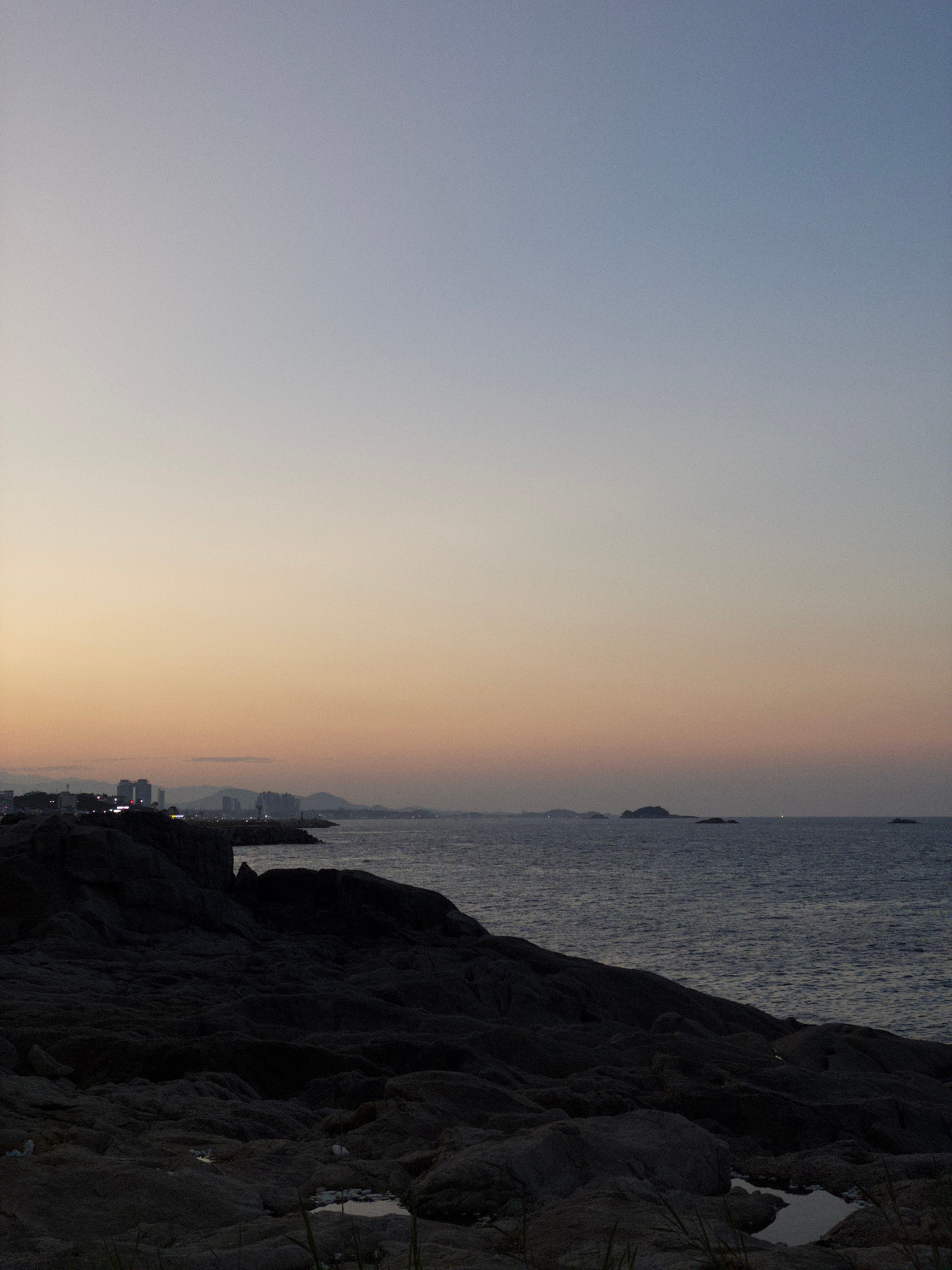 A rocky coastline at sunset, featuring a calm sea under a gradient sky transitioning from orange to blue