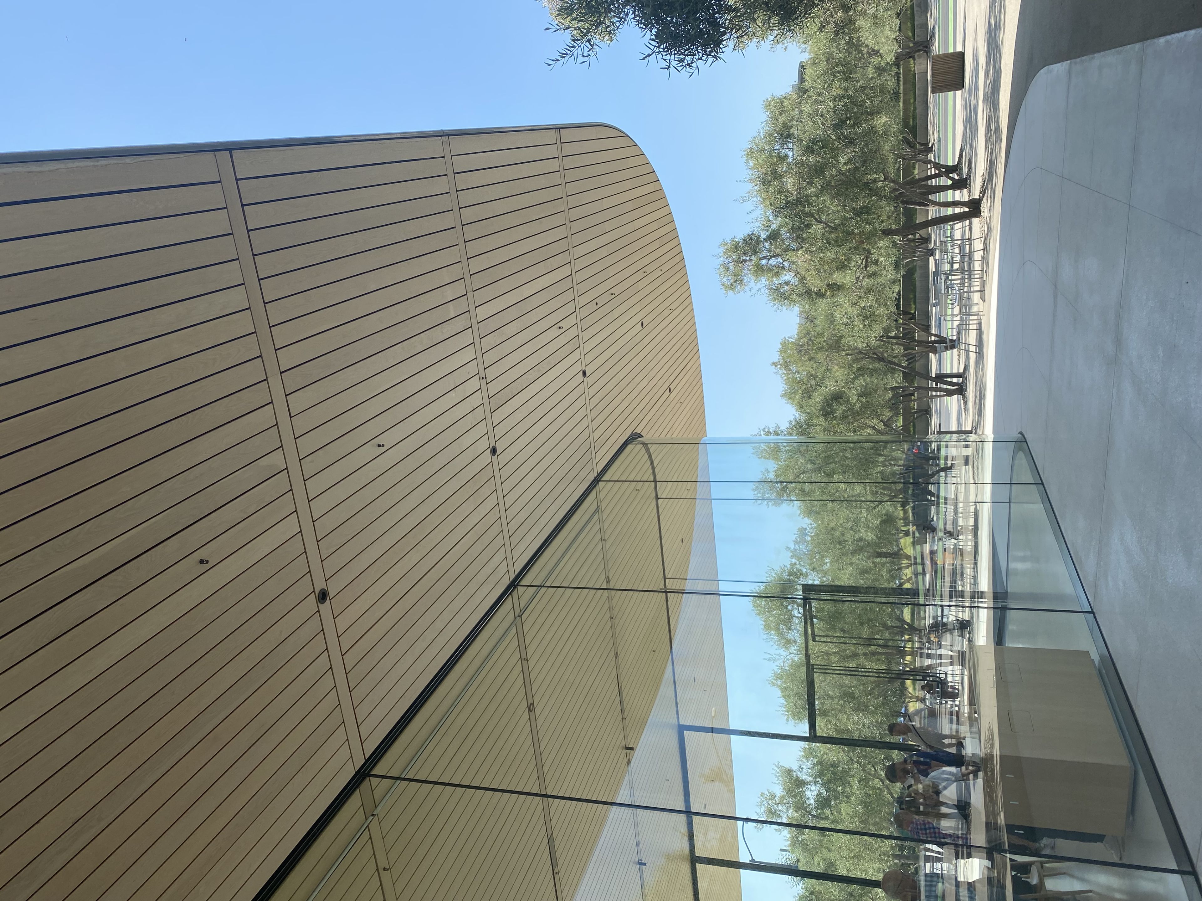A modern building with large glass walls and a wooden overhang, surrounded by trees under a clear blue sky