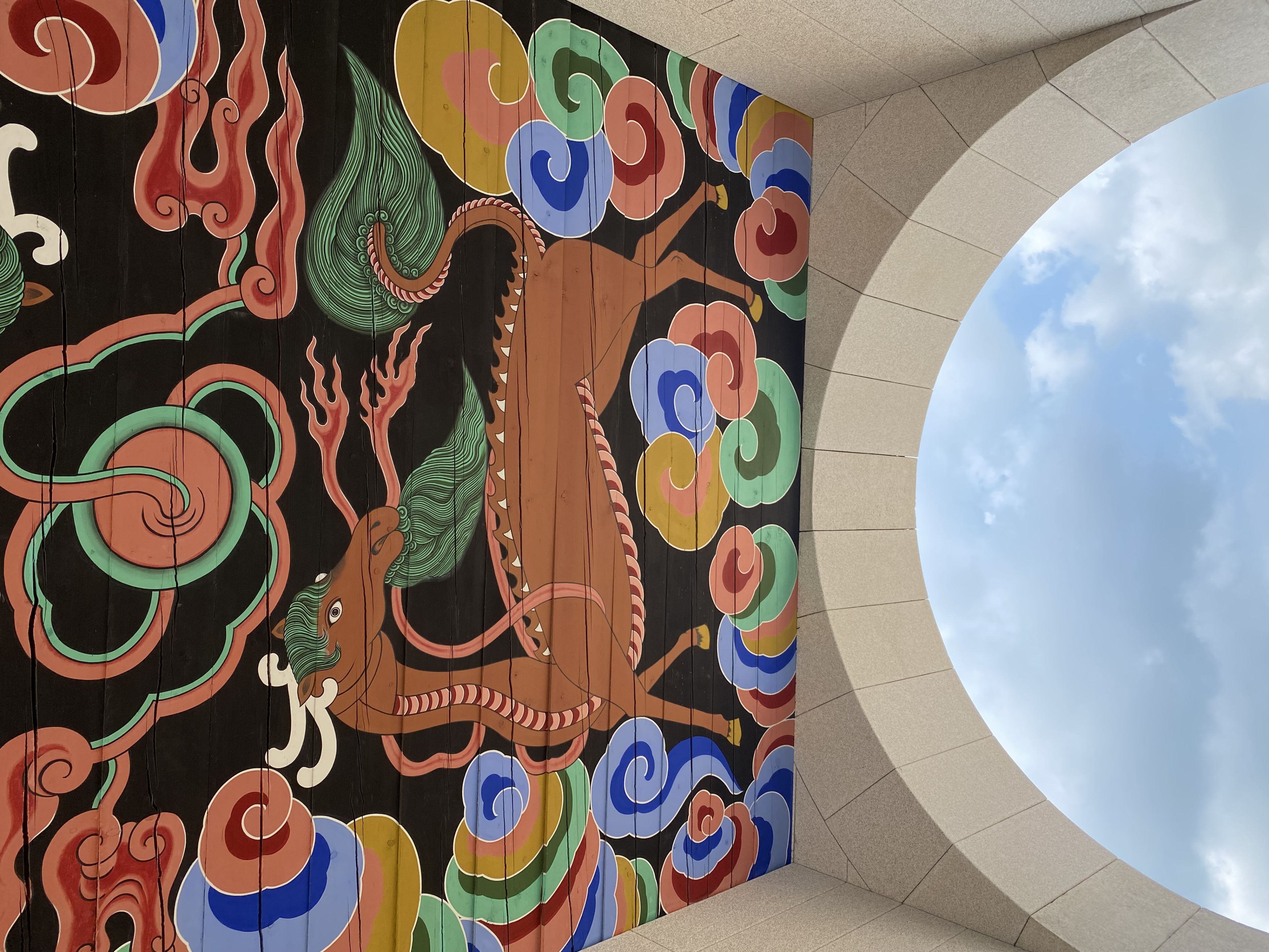 A colorful, ornate ceiling mural featuring a stylized dragon surrounded by swirling clouds and patterns, viewed through an archway with a glimpse of the sky