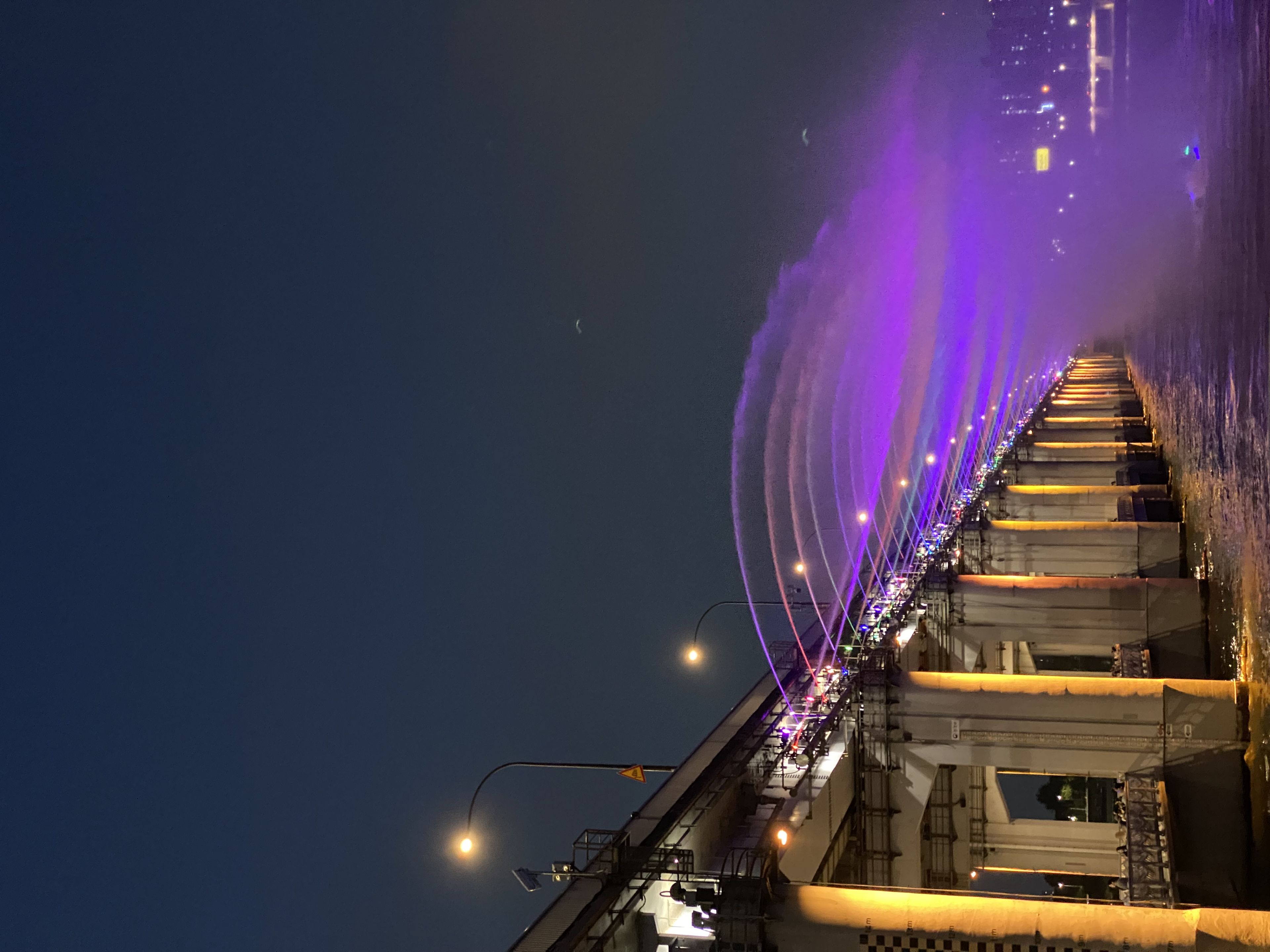A bridge at night with illuminated water jets creating a purple arc over the river