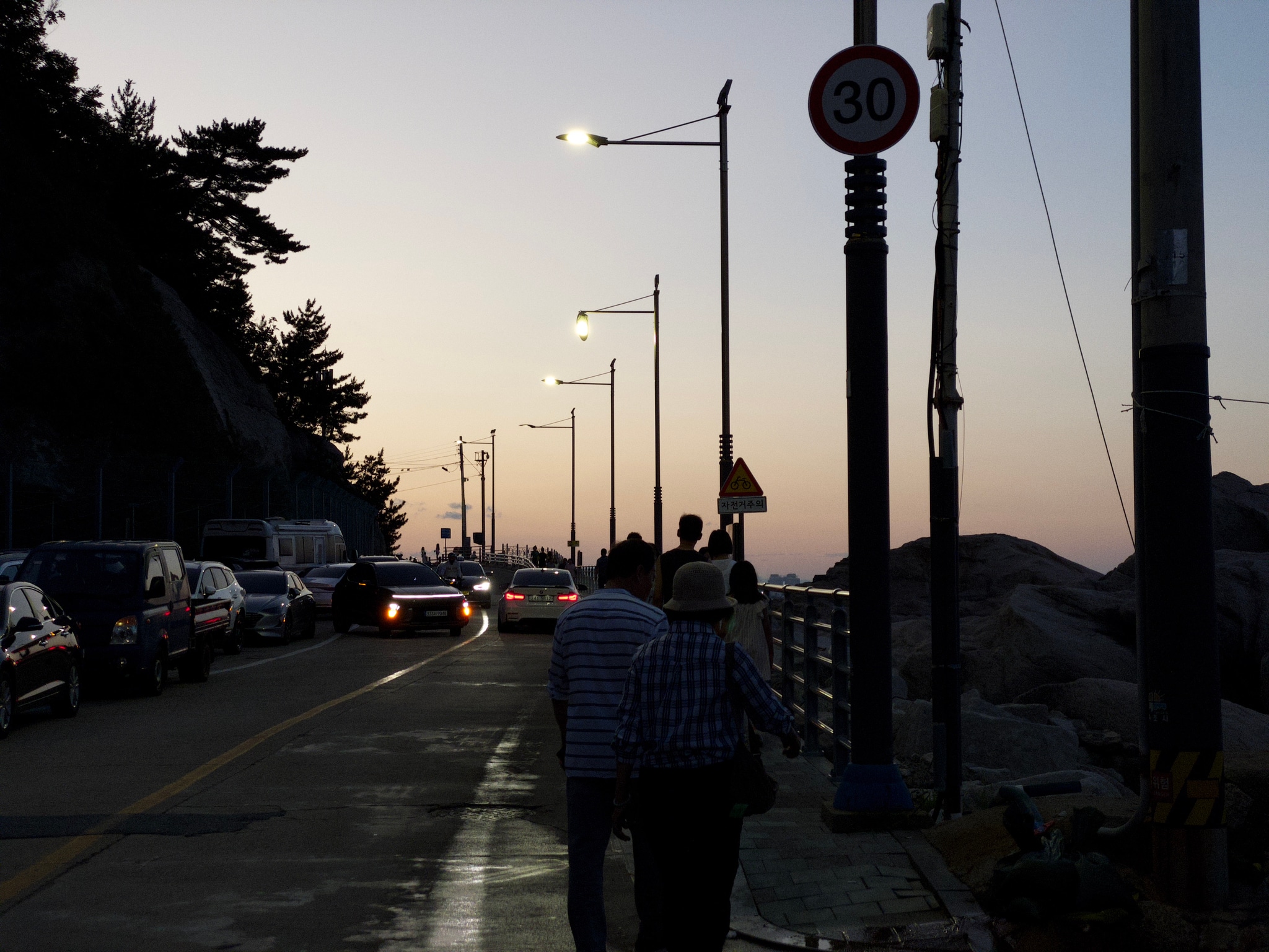 A seaside road at dusk with people walking, cars parked alongside, and streetlights illuminating the path. A 50 km/h speed limit sign is visible, and distant hills are silhouetted against the evening sky