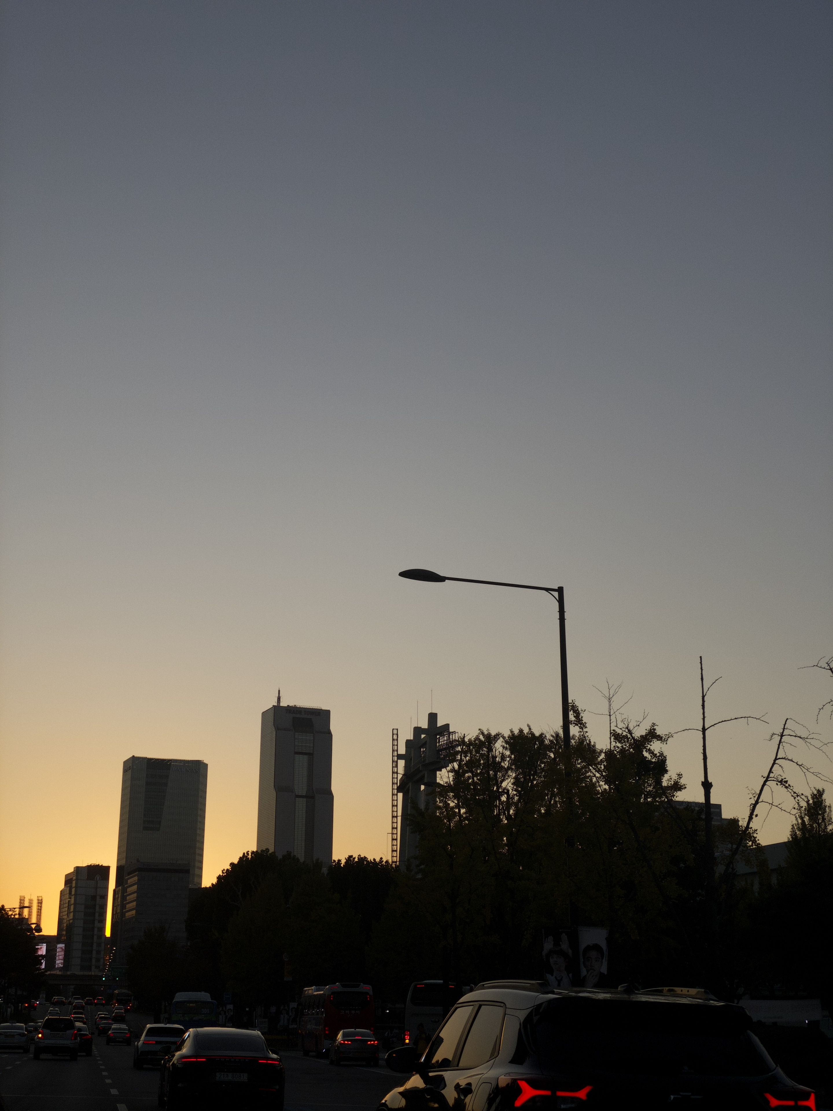 A cityscape at dusk with tall buildings silhouetted against a gradient sky, cars on the road, and a streetlamp in the foreground