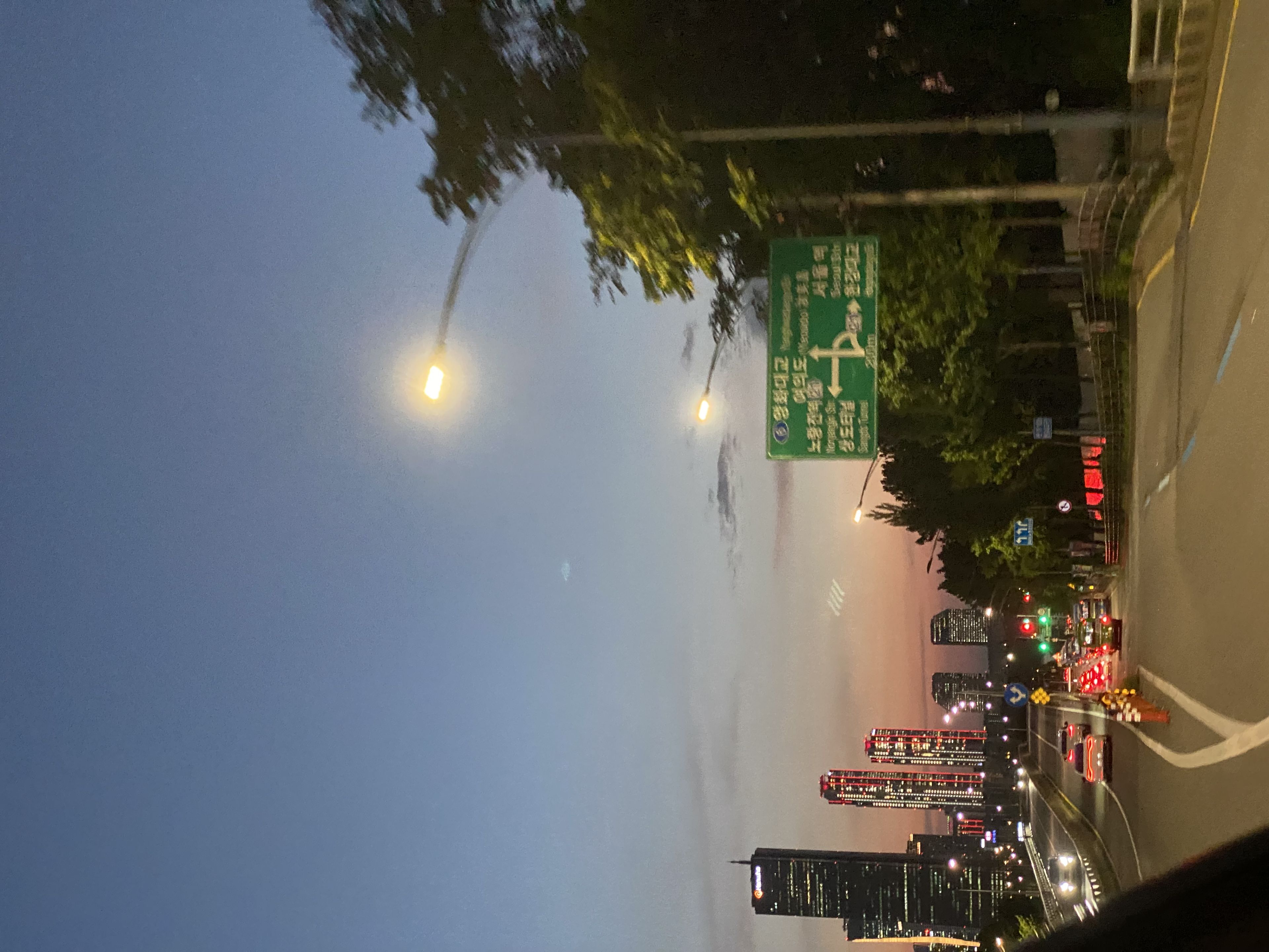 A cityscape at dusk with a road leading towards tall buildings, streetlights illuminating the scene, and a green road sign on the right
