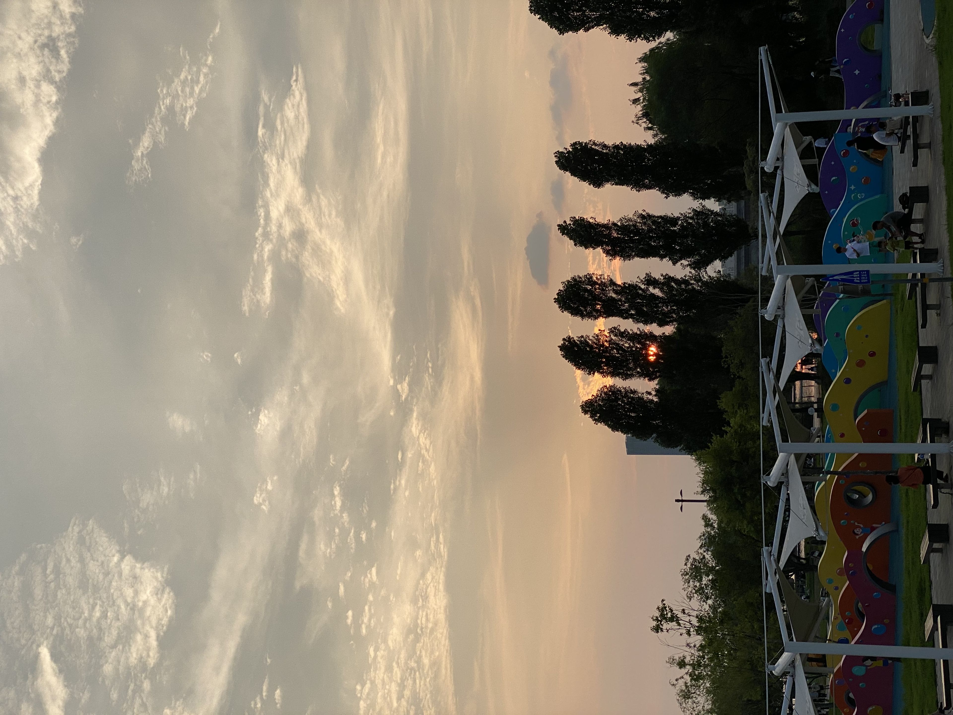 A sunset with a cloudy sky, tall trees silhouetted against the horizon, and colorful structures in the foreground