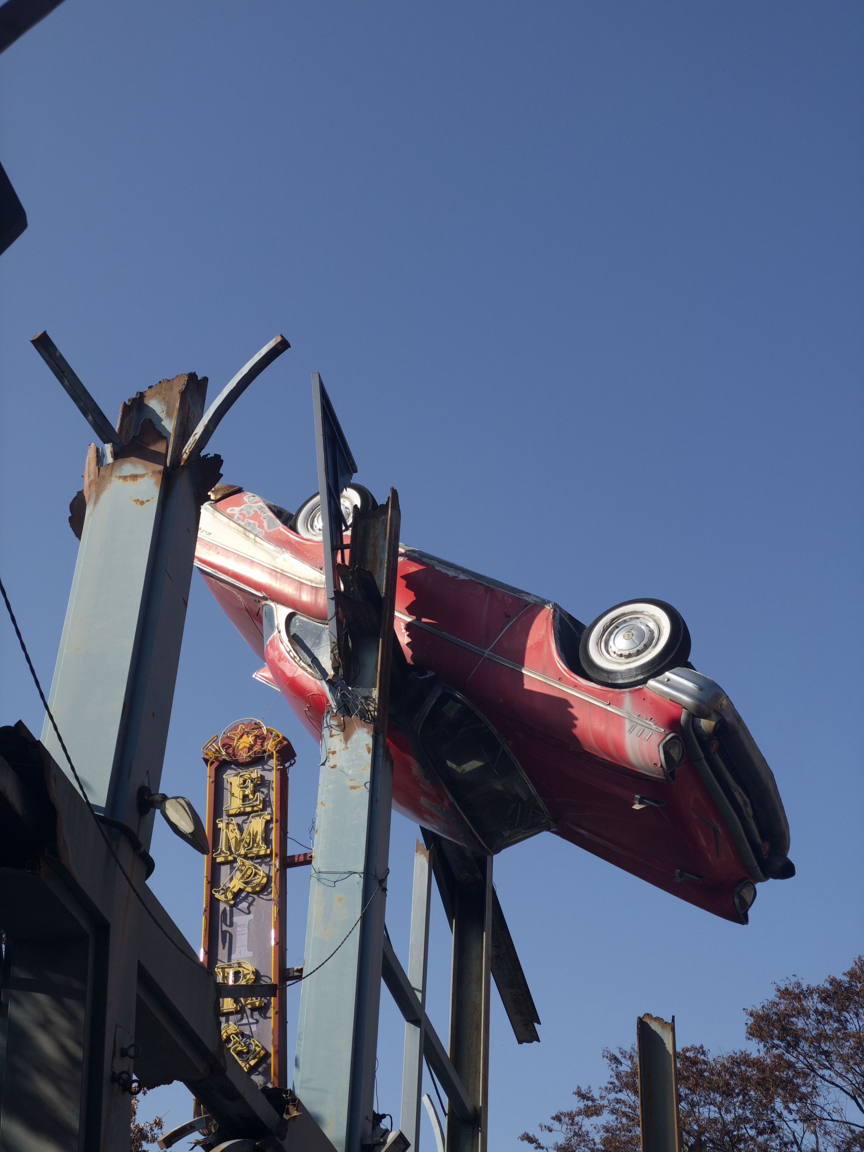 A red vintage car is mounted upside down on metal beams against a clear blue sky