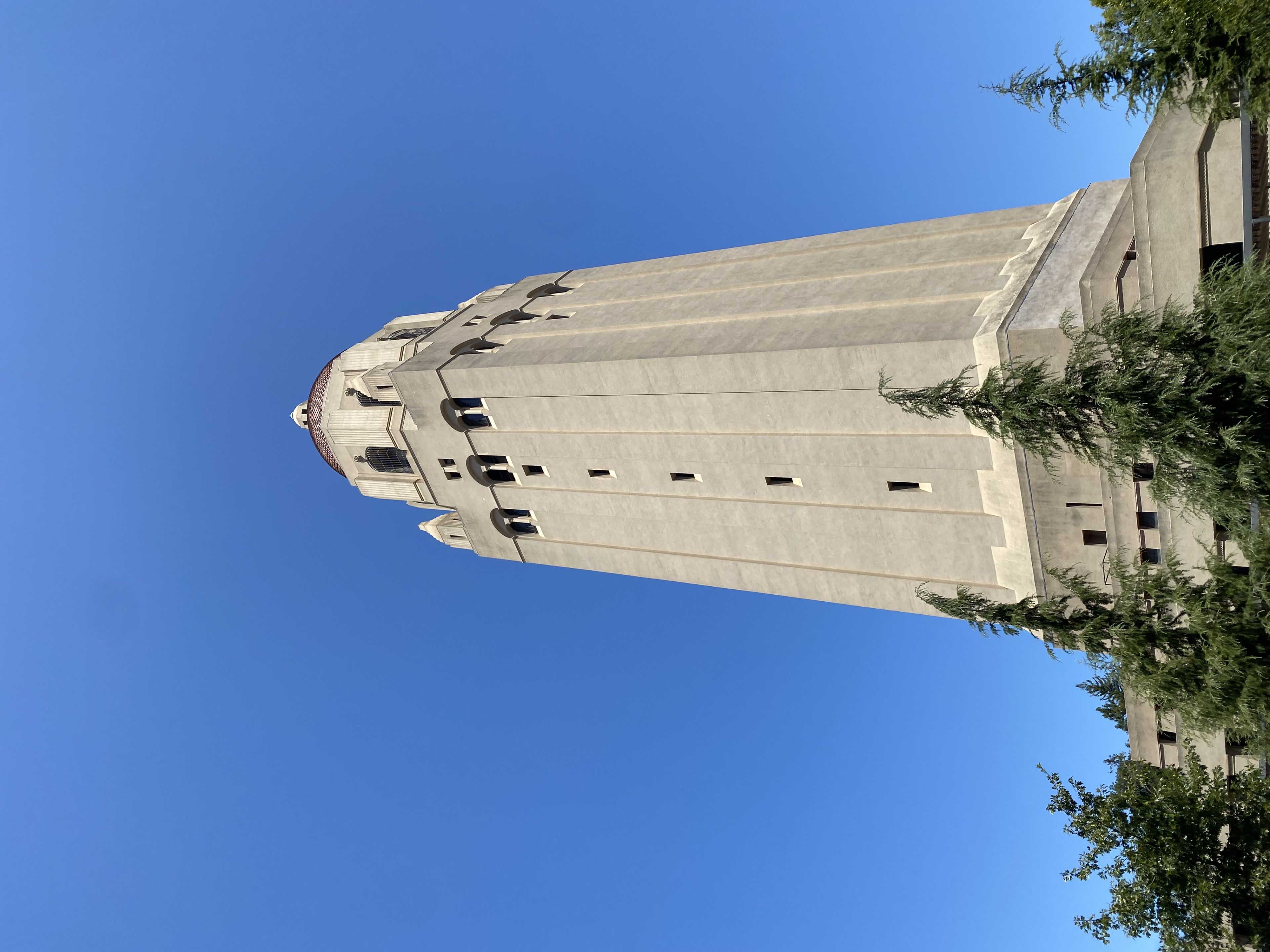 A tall, beige tower with a red-tiled roof set against a clear blue sky, surrounded by trees at the base